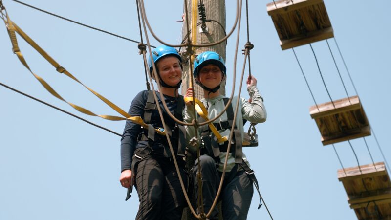 Two women wearing helmets and harnesses are suspended in the air, attached to ropes and cables. They appear to be on a high-ropes course or adventure activity, with wooden platforms visible in the background. The women are smiling, suggesting they are enjoying the experience. The sky is clear and blue, indicating a sunny day. The focus is on the subjects, highlighting their participation in the activity.